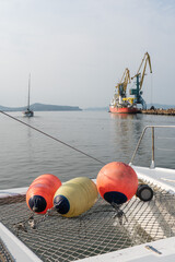 Colorful buoys rest on net at bow of catamaran with sailboat and port cranes beyond. Marine travel, sailing equipment, boat safety, nautical lifestyle, sea tourism, transportation. © Sofia
