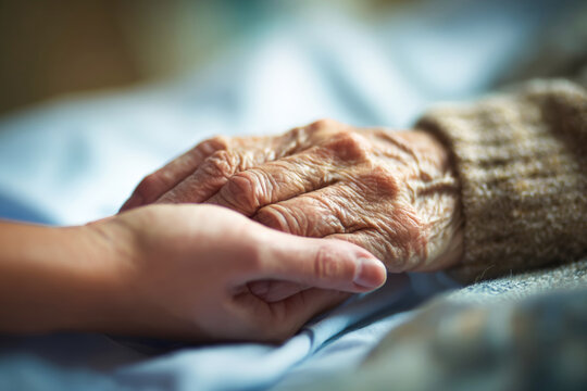 A nurse gently holds the hand of an elderly woman in a hospital room. The focus is on their hands, showing care and connection