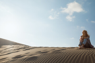 Fototapeta premium Young woman sits on soft sand dunes at Patara Beach, Turkey. Calm travel moment with warm sunlight, coastal scenery and relaxed lifestyle during summer vacation.