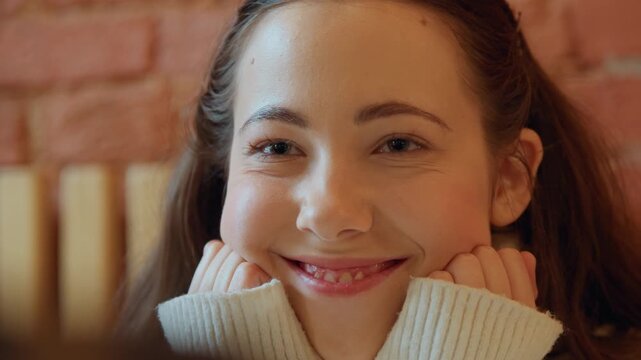 Close up of young woman with long brown hair wearing cream turtleneck sweater smiling and talking in front of brick wall. Her hands occasionally appear near her face as woman engages