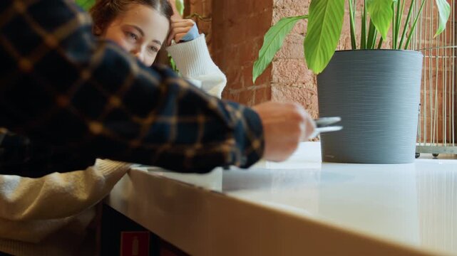 Close up of young woman using smartphone at white counter in cafe with brick wall and green plant. Another woman serves food on plate visible hands placing it near first subject who remains