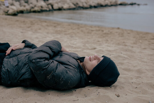 A teenager lies playfully on the sandy autumn beach, fooling around near the calm sea on a cold overcast day by the shore