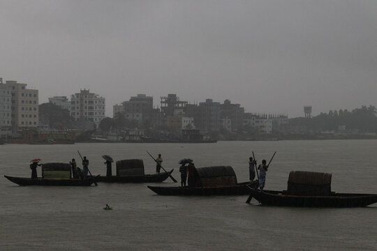 Narayanganj, Dhaka, Bangladesh - 13 July 2019: View of traditional wooden boats crossing the river with people holding umbrellas under an overcast sky.