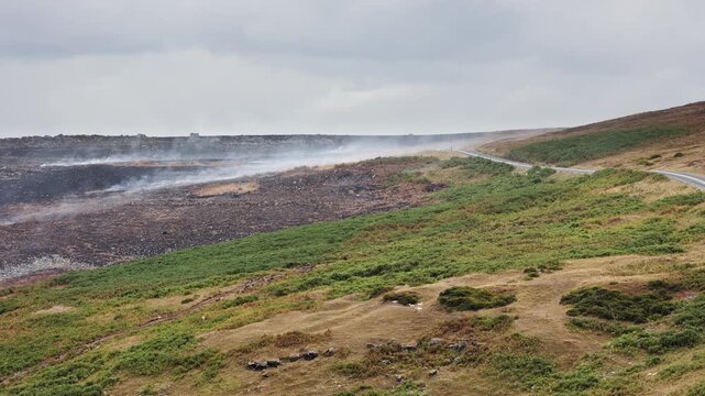 Smoke drifting across scorched earth after moorland fire, blackened ground beside green hillside and empty road, wide shot