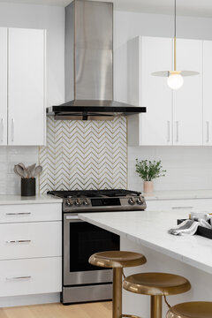 A white kitchen detail with a stainless steel range hood over a has range, a gold and marble chevron backsplash, subway tiles, and a marble countertop.