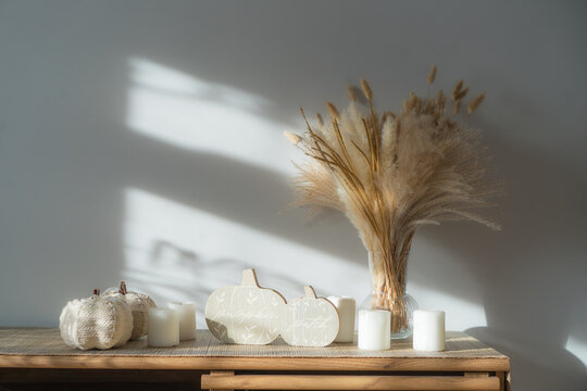 Cozy warm autumn home interior - white knitted pumpkins, wooden statuette, candles, dried flowers on the wooden console with white wall background. Scandinavian minimalist hygge home fall decor.