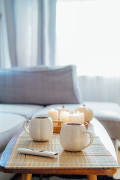 Autumn fall mood composition for hygge home decor. White pumpkin's shaped cups with hot drink, burning candles, remote controller on coffee table in living room. Movie night at home. Cozy relax time.