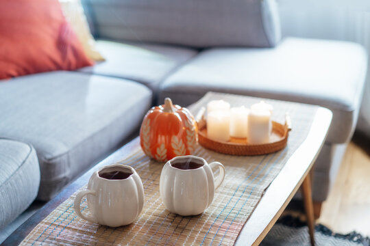 Autumn fall mood composition for hygge home decor. Hot tea or coffee drink in pumpkin's shaped cups, pumpkin decor and burning candles on the coffee table. Festive autumn, fall mood home decor