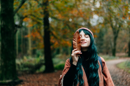 Portrait of Young smiling woman in hat partially covering her face with striking red fallen leaf enjoying autumn walk in forest. Feeling harmony, reunion with nature. Relaxing, personal fulfilment