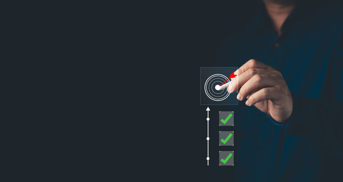 Businessman pointing at virtual target dartboard with green checkmarks. Achieve digital project goals, business strategy, success, and performance tracking concept on dark background.