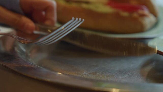 Close up of hand reaching for silverware on table with food in background. fork and knife rest on metal plates reflecting light softly as they remain stationary. scene features soft indoor