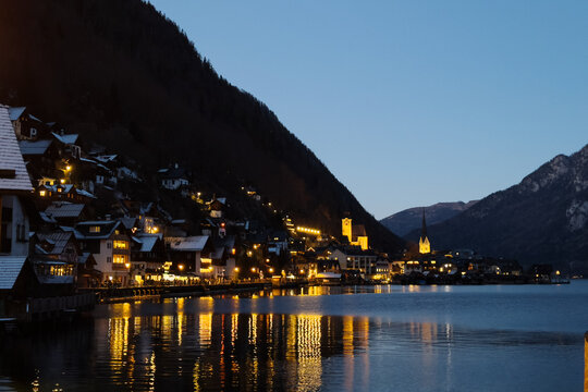 Beautiful night view of illuminated Hallstatt village in Austria reflected on calm lake water during dusk in winter. Iconic European landmark shot.
