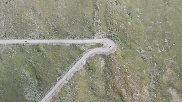 Cinematic overhead drone footage of two cars navigating a sharp hairpin turn on a remote Norwegian mountain road surrounded by rocky green landscape.