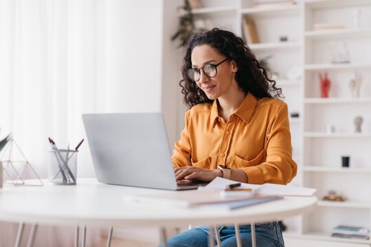Focused Asian Woman Working On Laptop At Home Office, Smiling While Typing On Computer, Sitting At Desk With Office Supplies. Engaged In Professional Work And Productivity. Business Concept.