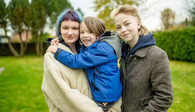 Three siblings pose together outdoors on a chilly autumn day, with a smiling boy hugging his older sister. Natural family portrait in a garden setting.