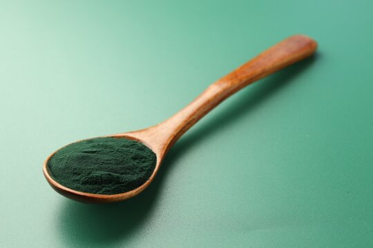 Natural spirulina powder in spoon on green table, closeup