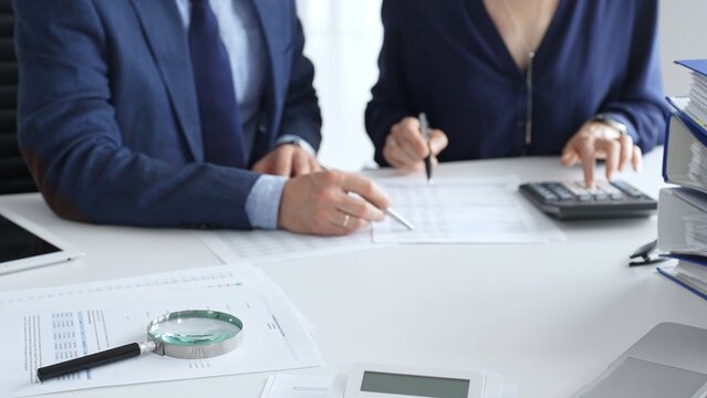 Close up of magnifying glass and calculator over financial documents opposite financial team, reviewing reports, collaborating on accounting details at modern office workspace. Audit and taxes