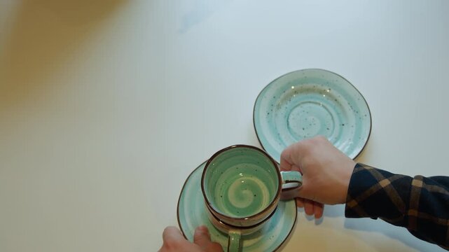 Close up of pair of hands carefully picking up matching ceramic cups and saucers with turquoise glaze from white table. sequence shows deliberate movement as each cup is lifted revealing