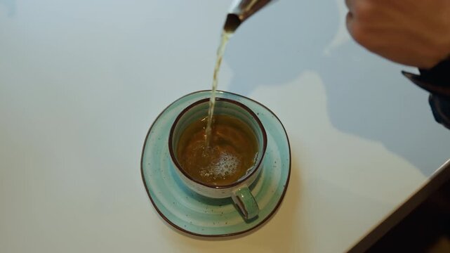 Close up of hand pouring tea into ceramic cup on saucer filling liquid and creating bubbles. scene features soft lighting with natural shadows cast by objects in focus emphasizing texture