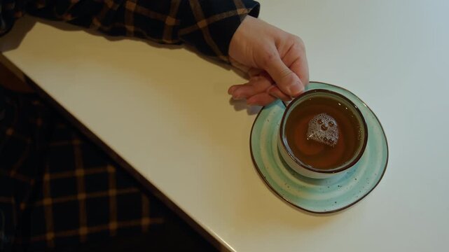 Close up of hand reaching for and lifting cup with tea on saucer from white table. Hand in plaid sleeve interacts gently revealing calm indoor setting with soft lighting and minimal