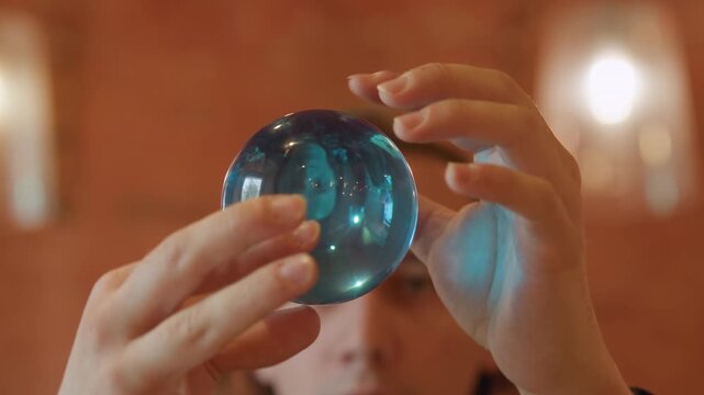 Close up of man in plaid shirt holding and rotating clear blue crystal ball examining its reflective surface. Hands manipulate sphere against warm indoor background with soft lighting