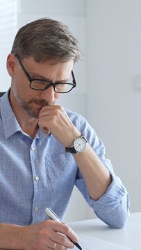 Businessman wearing blue sleek shirt, glasses, is thinking and concentrating while working at the desk in a bright, modern office, demonstrating professionalism and efficiency. Business people concept