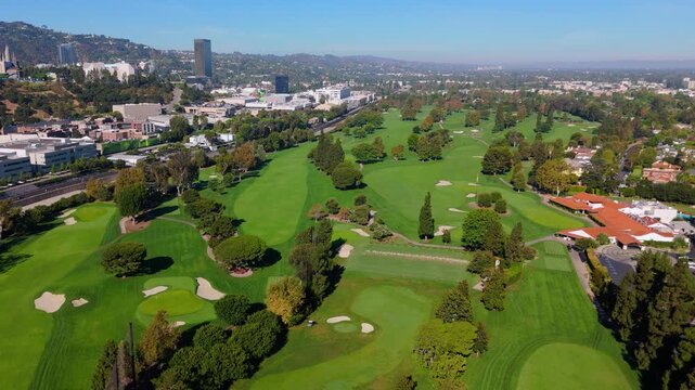 aerial view over manicured fairways, sand bunkers and mature trees of a golf course, with studio buildings, high-rise towers and theme park structures of Universal City rising beyond, and the