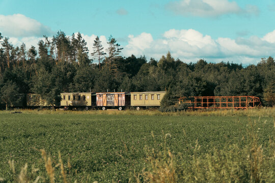 A line of old green vintage railway carriages and a rusty flatcar stands near a green forest and a field under a blue sky with white clouds.