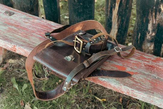 A vintage dark brown leather shoulder bag with straps and metal buckles rests on a weathered red and grey wooden bench outdoors.