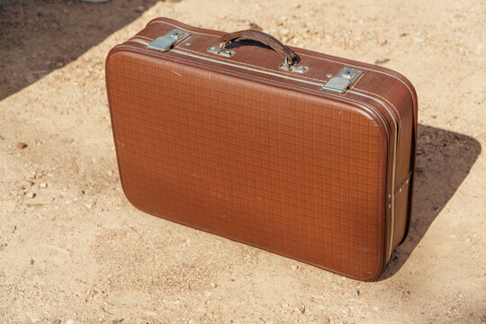 A structured brown vintage suitcase with a checkered texture and metal clasps stands on a sandy dirt ground outdoors.