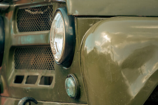 A close-up view of the front section of a vintage olive green military vehicle showing its round headlight, grille, and glossy fender.