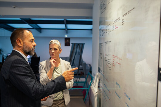 Two business colleagues discussing strategy at glass whiteboard in modern office
