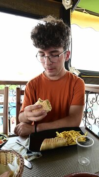 Marrakech, morocco. August 8, 2026. Young man with curly hair and glasses having lunch at an outdoor restaurant in marrakech, holding a traditional moroccan kebab wrap