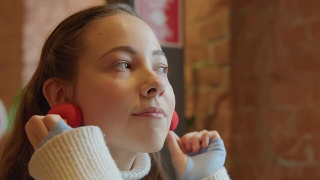 Close up of woman holding red spherical objects near her face in indoor setting with soft lighting. Woman wears white turtleneck sweater and background features warm tones like brick walls
