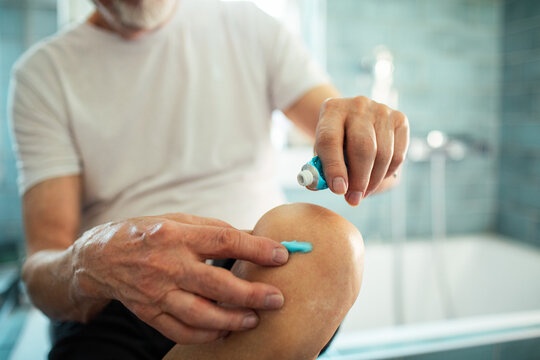 Senior man applying topical pain relief gel to sore knee in home bathroom