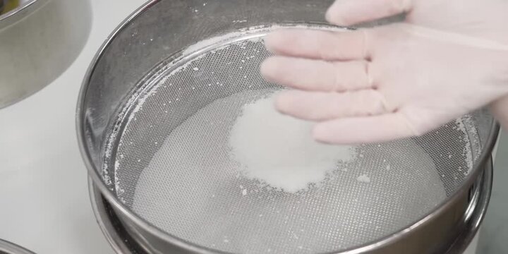 close-up shot of a person wearing protective gloves pouring a fine white powdery substance from a small metal bowl into a large stainless steel mesh sieve. The fine powder gathers in the center of the