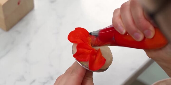 A professional pastry chef or baker uses a plastic piping bag with a petal tip to create an elegant red frosting flower on a metal decorating nail. This detailed, high-angle shot captures the precisio