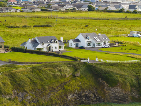 Aerial view of white houses situated on a grassy cliff edge overlooking the coastline with green fields and a residential neighborhood in the background in Bundoran, County Donegal, Ireland.