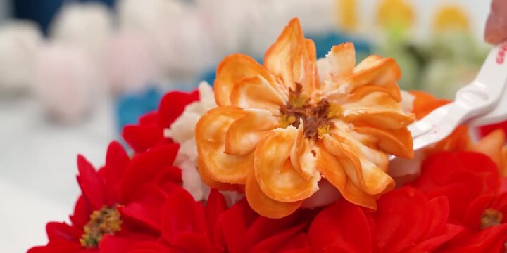 A vibrant, detailed close-up shot capturing a skilled confectioner's work. It highlights an intricately piped orange marigold flower resting atop a bed of rich red buttercream blossoms. The shallow de
