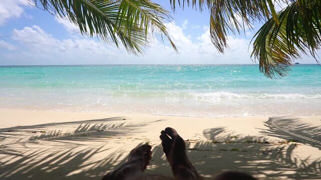 A person rests feet up on a sandy tropical beach in Maldives under palm tree shade, looking out at turquoise ocean waves on a sunny day.