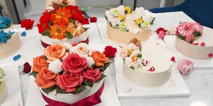 A high-angle shot showcasing a collection of artisanal celebration cakes meticulously decorated with vibrant buttercream flowers. The display features a variety of intricate piping styles, including r