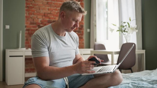 Casual man sitting on bed enters payment details on his mobile phone in bright bedroom interior
