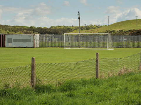 White soccer goal stands alone on green field. Cloudy sky above, hills in background. Scene quiet, empty, peaceful. Ideal for sports, training, or rural themes. Big game preparations.