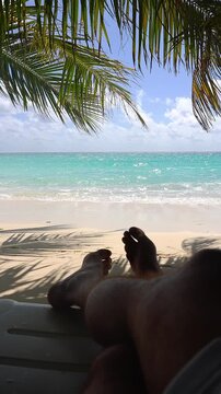 A person rests on a sunbed under palm fronds looking out at turquoise ocean waves crashing onto a sandy shore in Maldives during sunny weather.