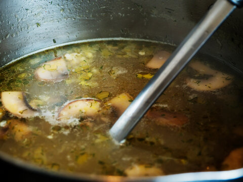 Mushroom soup simmers in stainless steel pot on black stovetop. Wooden surface nearby adds rustic feel. Warm, inviting kitchen scene perfect for recipes or food blogs. Ideal for cooking, home