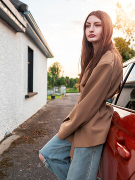 Young woman leans against red car beside textured wall. Warm golden light bathes scene in soft glow. She gazes calmly ahead with relaxed posture. Suitable for travel, lifestyle, or automotive themes