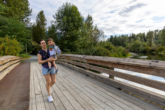 Mother Carrying Young Son Walking Across Wooden Bridge In Great Blue Heron Nature Reserve, Chilliwack BC Canada
