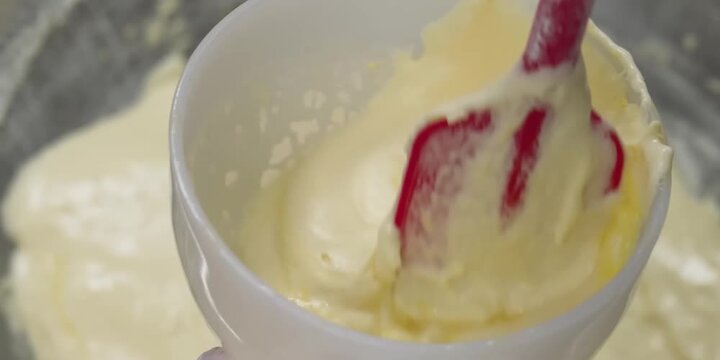 Close-up shot of a pastry chef carefully tempering a raw egg yolk inside a small white ceramic bowl by folding in a warm, creamy batter or custard base. This essential baking technique prevents the eg