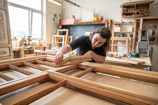 Attentive female carpenter carefully examines parts of antique wooden window frame before complete restoration process. professional craftswoman holds part of window and inspecting quality of wood.