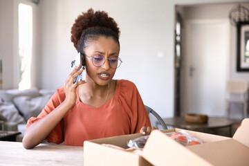 Worried African woman making phone call while inspecting package with incorrect or damaged product,...